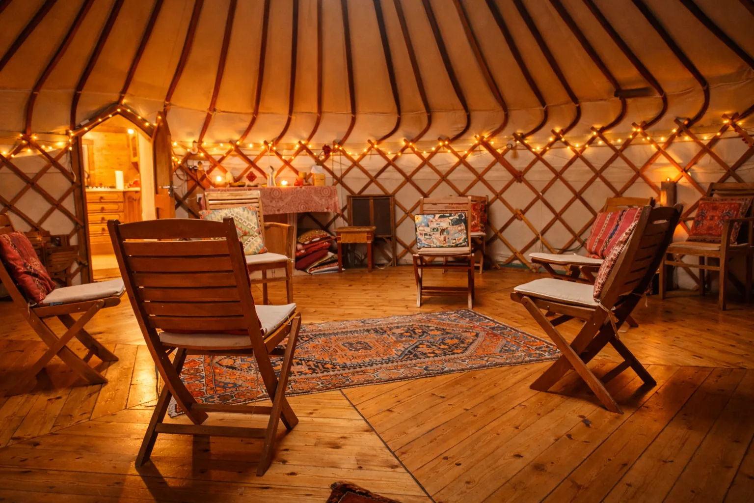 Inside the yurt, warm fairy lights, wooden chairs arranged in a circle with cushions and a rug